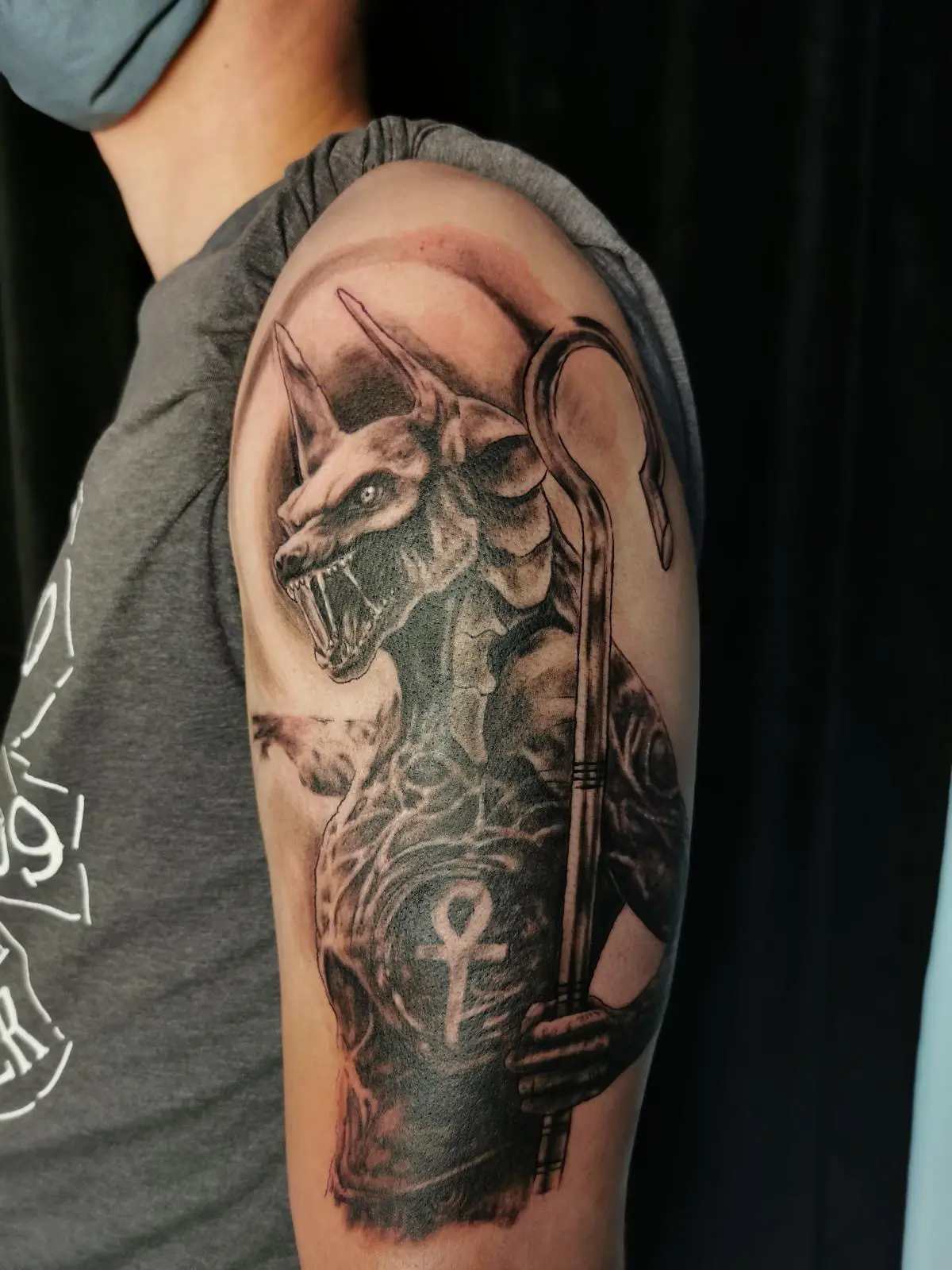Realistisches Black-&-Grey-Tattoo von Anubis mit Stab und Ankhsymbol auf dem Oberarm