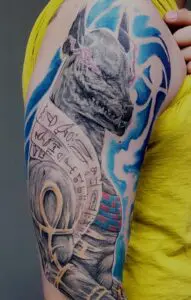 Farbiges Anubis-Tattoo auf dem Oberarm mit ägyptischen Symbolen und blauem Hintergrund