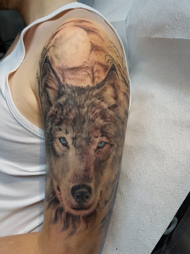 Ein realistisches Tattoo eines Wolfskopfes mit leuchtend blauen Augen auf dem Oberarm. Wolf-Tattoo von Ozzy in Hamburg gestochen.